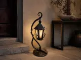 Kuta lampa stojąca umieszczona na kamiennej posadzce w rustykalnym wnętrzu, emitująca ciepłe, rozproszone światło przez matowy klosz.